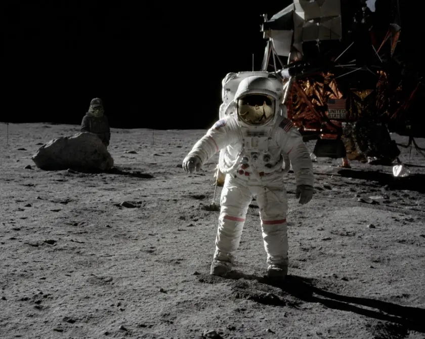 An astronaut beside the lunar lander, with a small green figure peeking from behind a boulder